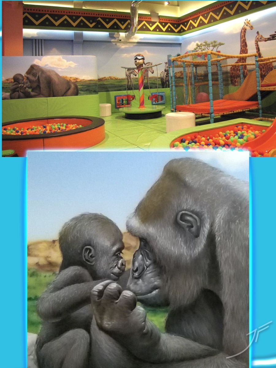Zona de juegos infantil con mural de sabana, jirafas, cebras y una gorila con su cría, junto a piscinas de bolas